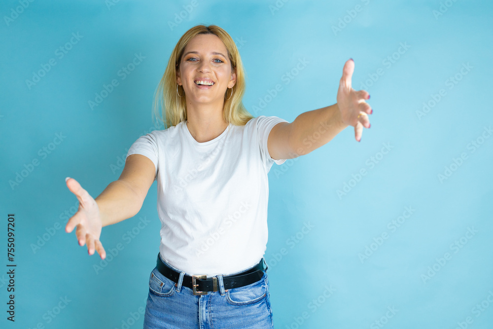 Young beautiful woman wearing casual t-shirt over isolated blue background looking at the camera smiling with open arms for hug Young beautiful woman wearing casual t-shirt over isolated blue background looking at the camera smiling with open arms for hug