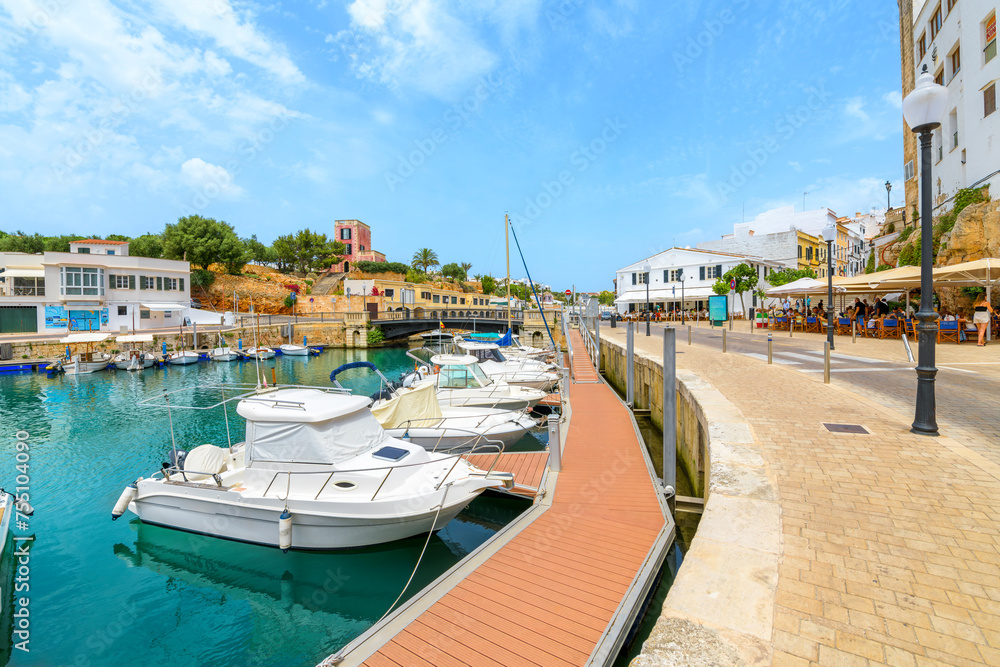 Boats line the picturesque marina port harbor full of shops and sidewalk cafes at the seaside town of Ciutadella de Menorca, on the Balearic island of Menorca in the Mediterranean Sea.	