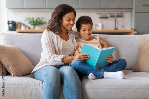 Happy little black boy reading book with his mom at home