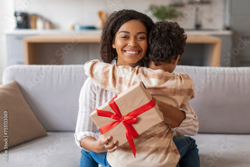Happy black mother embracing little son and holding gift with red ribbon
