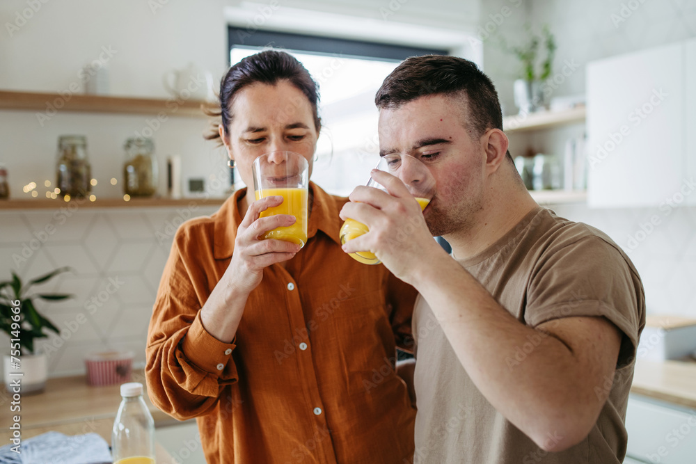 Fototapeta premium Portrait of young man with Down syndrome with his mother at home, toasting, drinking juice. Morning routine for man with Down syndrome.