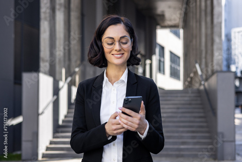 Close-up photo of a smiling young business woman in a suit and glasses standing outside an office building and using a mobile phone