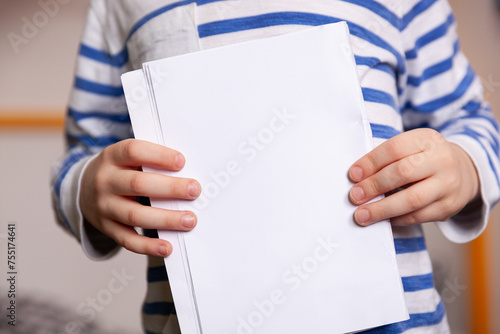 A closeup kid boy in a striped jacket holds white sheets of paper in his hands.
