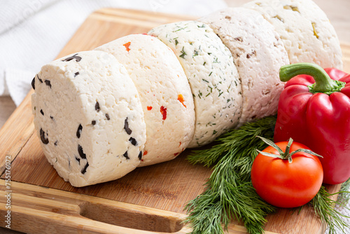 Closeup of several varieties of homemade cheese with different fillings with paprika, dill, olives, walnuts and cookies on a wooden board with fresh dill, tomatoes and bell pepper.