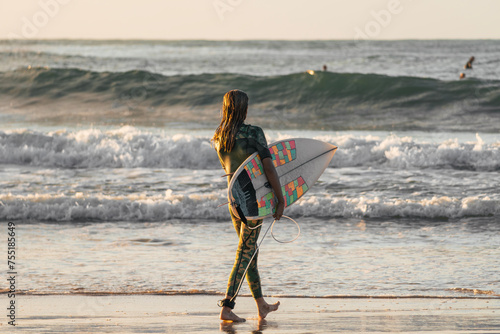 Young surfer girl in camo wetsuit entering the sea with her surfboard held in one arm
