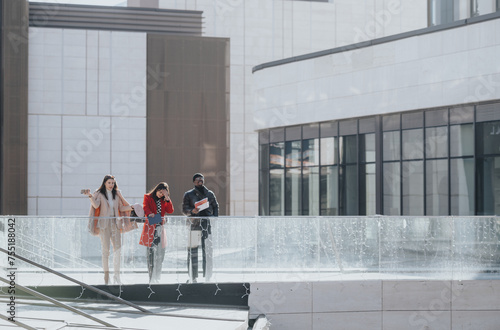 Three young adults casually dressed in urban fashion, walk and converse beside a glass railing with a modern building background. © qunica.com