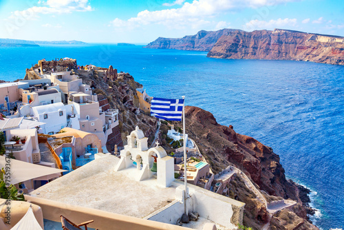 Greek orthodox church with bells, greek flag and famous white houses on Santorini island, Aegean sea, Greece.