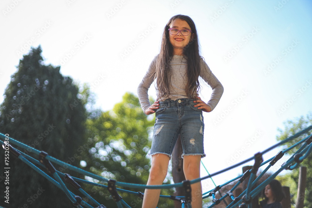 Obraz premium A caucasian girl of eight years old stands on a rope swing with a happy smile in the park