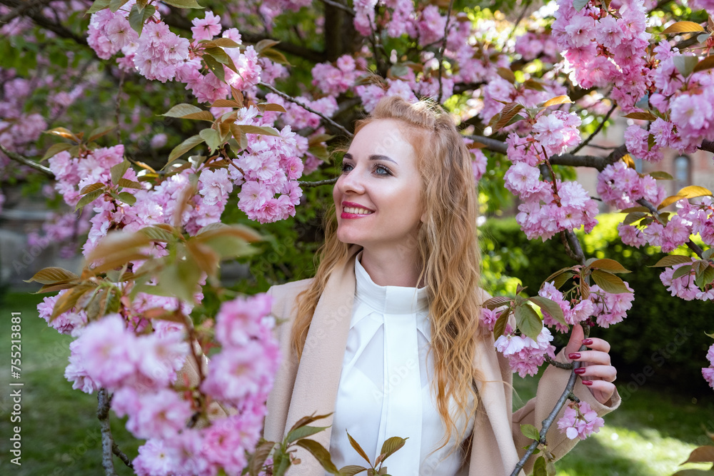 Obraz premium Redhead girl in white shirt stands near sakura in sunny spring weather. Portrait of attractive woman with wavy hair in blooming park.