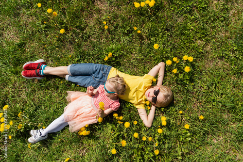 Children boy and younger sister are lying on green field with dandelions. Top view. Siblings rest in the grass.