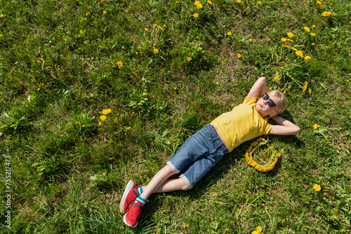 Boy in yellow t-shirt and jeans shorts with hands behind head lying on grass and rest. Countryside. Top view.