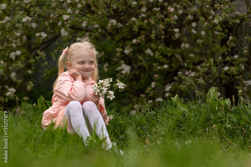 Little smiling blonde girl leans her cheek against her fist, holds bouquet of flowers and sits on grass. Blooming trees, spring garden