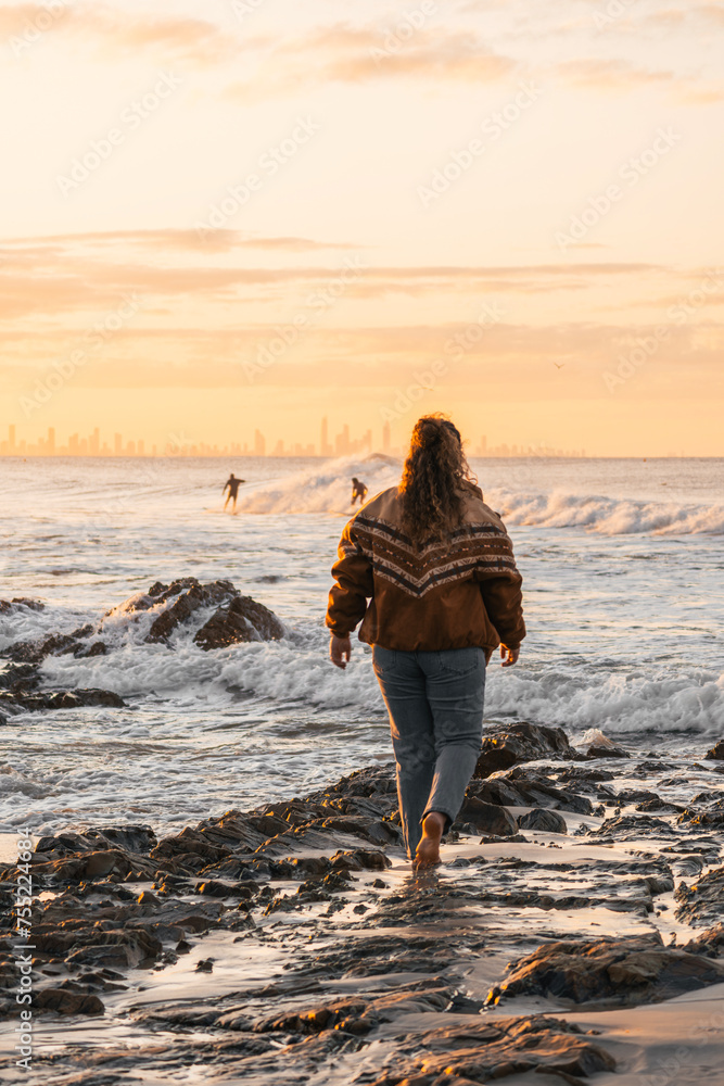 Girl walking on beach rocks barefoot while watching surfers during sunset Girl walking on beach rocks barefoot while watching surfers during sunset