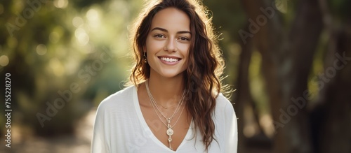 A young woman with long hair is smiling while wearing a white shirt and a pearl necklace. She is looking directly at the camera, with blurred trees in the background.