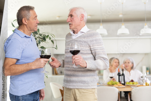 During friendly meeting, two elderly men with glass of wine in their hands stand at distance from table, communicate and chatting, discussing sports news and hobby