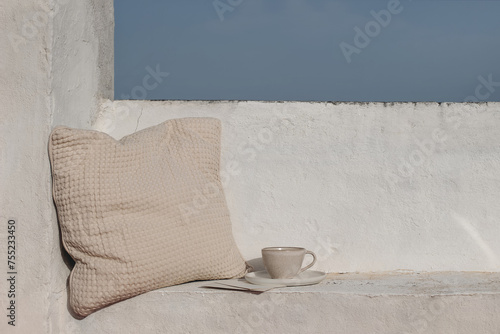 Summer vacation still life, relaxation concept. Mediterranean outdoor sitting area. Bench with beige waffle textured cushion. Cup of coffee. White wall background. Blue sky, travel banner