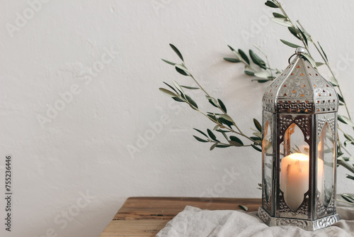 Ramadan Kareem, Eid ul Fitr holiday still life. Ornamental silver Moroccan lantern with olive tree branches on wooden table, bench. Burning candle. Blurred white wall background. Iftar decor.