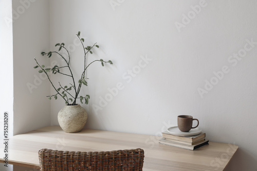 Scandinavian living room interior. Cup of coffee, books on wooden table. Willow tree branches, catkins in vase. Rattan chair. Elegant Scandi working space, home office. Blank beige wall background