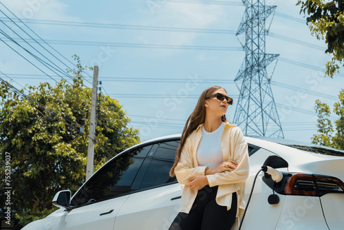 Young woman recharge EV car battery at charging station connected to power grid tower electrical industrial facility as electrical industry for eco friendly vehicle utilization. Expedient photo