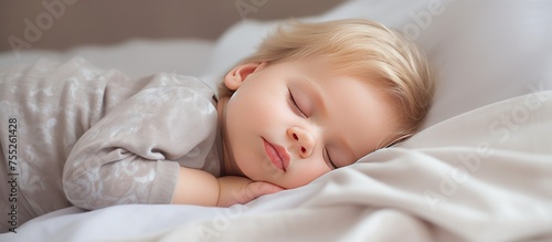A white, blond Caucasian baby toddler is peacefully sleeping on a bed indoors. The child is lying down, with eyes closed, deep in sleep. The toddler appears cute and adorable.