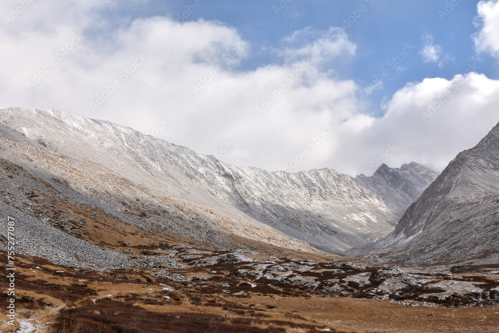The steep slopes of a high mountain range descend into a picturesque valley sprinkled with the first snow under a cloudy autumn sky.