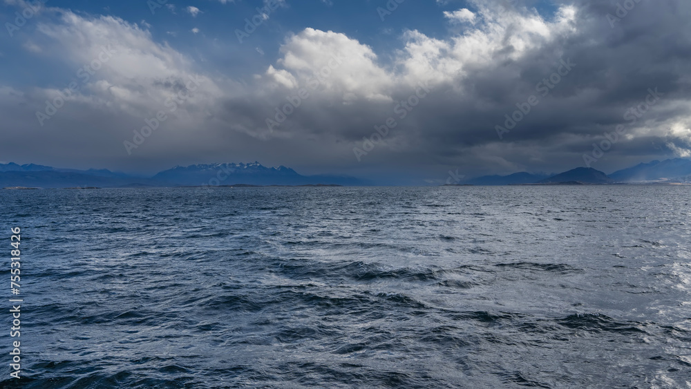 The surface of the blue ocean with small waves and ripples. In the distance, the Andes mountain range is visible against the sky and clouds. The Beagle Channel. Argentina. Tierra del Fuego Archipelago