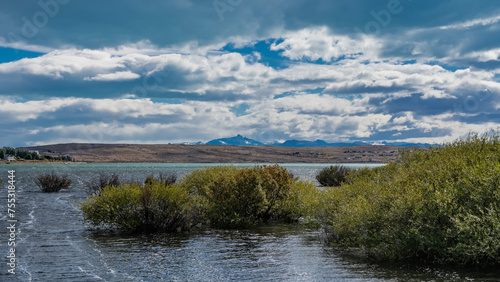 Lush green bushes grow near the shore of the blue-turquoise lake. Ripples on the water. Hills and mountains against the sky and clouds in the distance. Lago Argentino. Redonda Bay. El Calafate. 