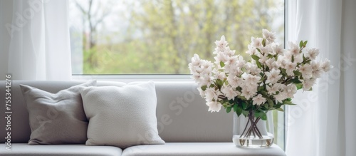 A soft gray couch with cushions is positioned next to a window adorned with a bouquet of white flowers in a modern living room, alongside a television.