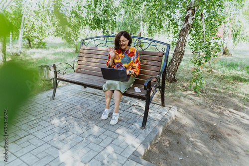 Outdoor office with a cheerful woman conducting video meetings on her computer in nature. Cheerful woman engages in lively video calls on her laptop while sitting on park bench
