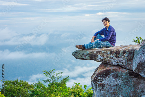 A handsome Asian man is sitting on a large rock that juts out from a steep cliff named Sut Phaen Din. Located at Pa Hin Ngam National Park in Thep Sathit district, Chaiyaphum province, Thailand. photo