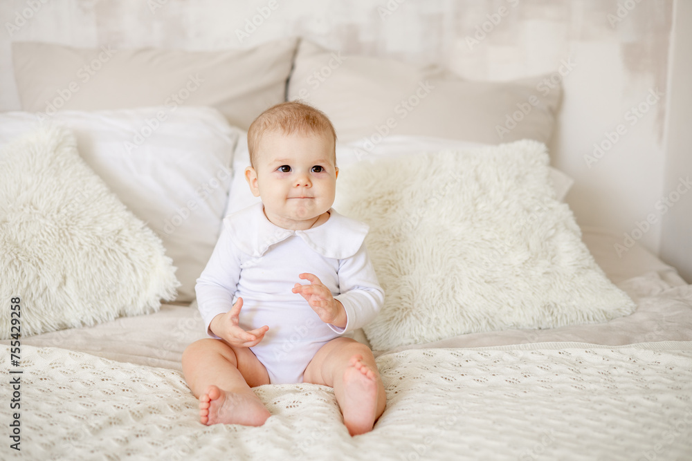 portrait of a little baby girl of six months on a bed at home in a bright bedroom smiling, happy newborn in a white cotton bodysuit portrait of a little baby girl of six months on a bed at home in a bright bedroom smiling, happy newborn in a white cotton bodysuit