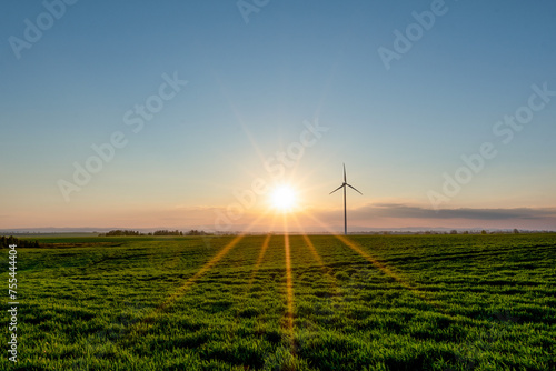 Sunset with sunrays over a green field with a wind turbine in the foreground