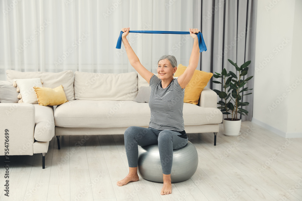 Fototapeta premium Senior woman doing exercise with elastic resistance band on fitness ball at home