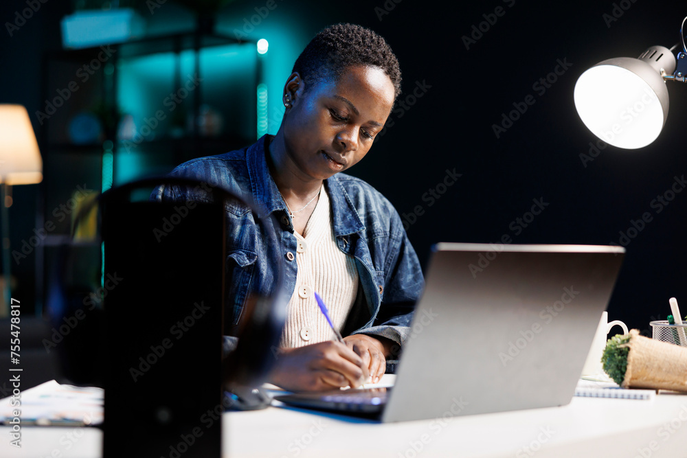 African American woman works diligently at her desk using her laptop and pen, researching, and brainstorming ideas. A productive and focused individual utilizing technology for work and study. African American woman works diligently at her desk using her laptop and pen, researching, and brainstorming ideas. A productive and focused individual utilizing technology for work and study.