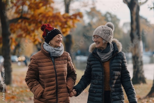 Two women are walking together in the park, both wearing hats and warm coats. They are smiling and seem to be enjoying each other's company