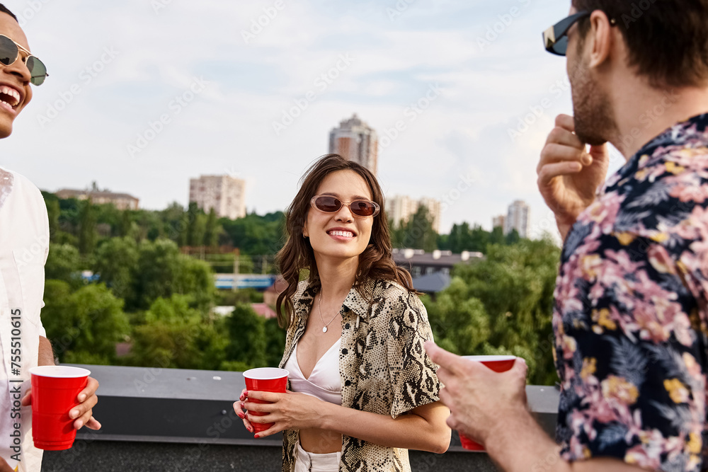 joyous interracial friends in casual attire with sunglasses drinking from red cups on rooftop