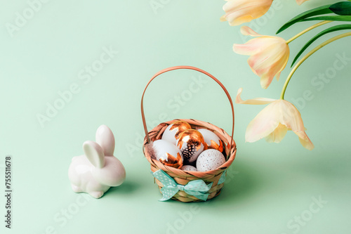 Basket with painted white and golden eggs, bunny and yellow tulips on light green background. Easter concept. Closeup #755511678