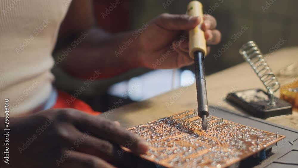 Cropped shot of unrecognizable Black male engineer in orange coverall pants and white undershirt soldering PCB at table in workshop Cropped shot of unrecognizable Black male engineer in orange coverall pants and white undershirt soldering PCB at table in workshop