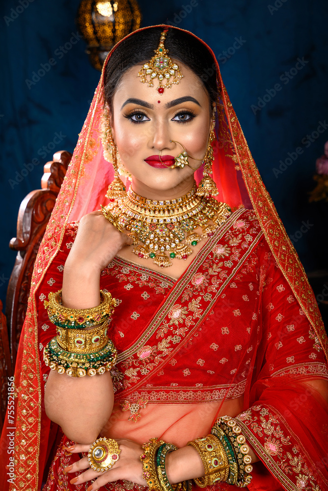 Stunning Indian bride dressed in traditional red bridal lehenga with heavy gold jewellery and veil sitting in a chair smiles tenderly in studio lighting. Wedding fashion and lifestyle.