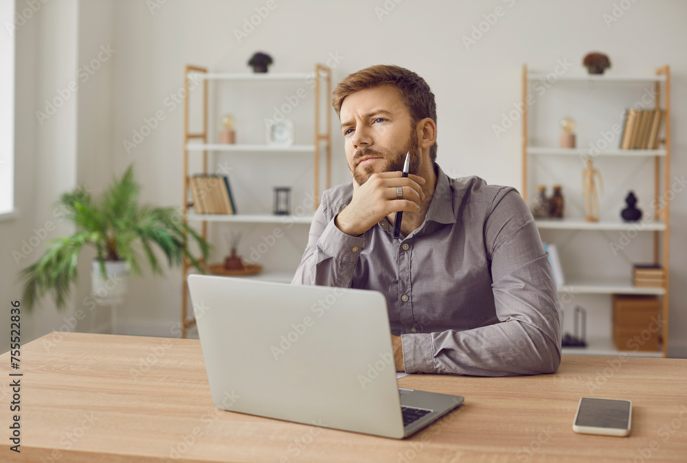 Pensive writer working in his home office. Male novelist thinking of plot of his new book. Young man sitting at desk with laptop computer, holding hand on chin, looking away, and thinking Pensive writer working in his home office. Male novelist thinking of plot of his new book. Young man sitting at desk with laptop computer, holding hand on chin, looking away, and thinking