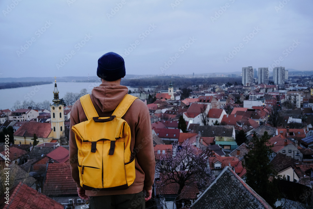 Danube river, tiled red roofs of houses and clock tower of church. A young guy backpacker travels and enjoys tourist spots. Belgrade, Serbia the old town Zemun. Rear view. Danube river, tiled red roofs of houses and clock tower of church. A young guy backpacker travels and enjoys tourist spots. Belgrade, Serbia the old town Zemun. Rear view.