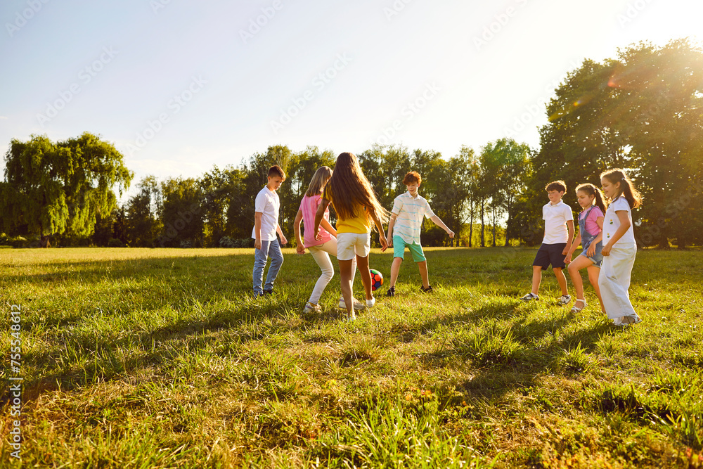 Obraz premium Happy children boys and girls running towards ball playing together in the park on the green grass on a summer holidays. Teenage laughing kids in casual clothes playing football in the garden.