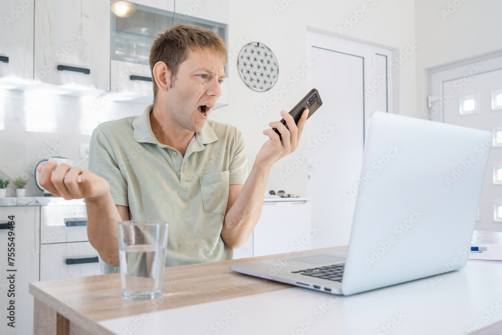 Angry male arguing and screaming on mobile phone while working on laptop online at home. Annoyed man freelancer shouting into the phone gesturing, describing a problem to be solved. Angry excited guy