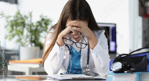 Tired woman doctor sitting at table and holding glasses in clinic. Treatment of critical patients concept photo