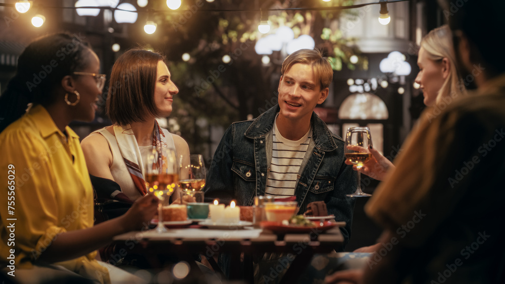 Diverse Group of Friends Enjoying Nightlife in a Street Cafe. Young Women and Men Sitting Behind a Table, Having Fun and Joyful Conversations. Caucasian Man Sharing Entertaining Life Stories
