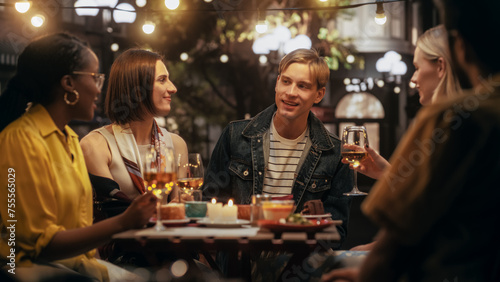 Diverse Group of Friends Enjoying Nightlife in a Street Cafe. Young Women and Men Sitting Behind a Table, Having Fun and Joyful Conversations. Caucasian Man Sharing Entertaining Life Stories