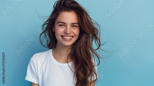 Pretty young smiling woman with dark hair on a blue background with copy space. Cute girl in a simple white T-shirt.