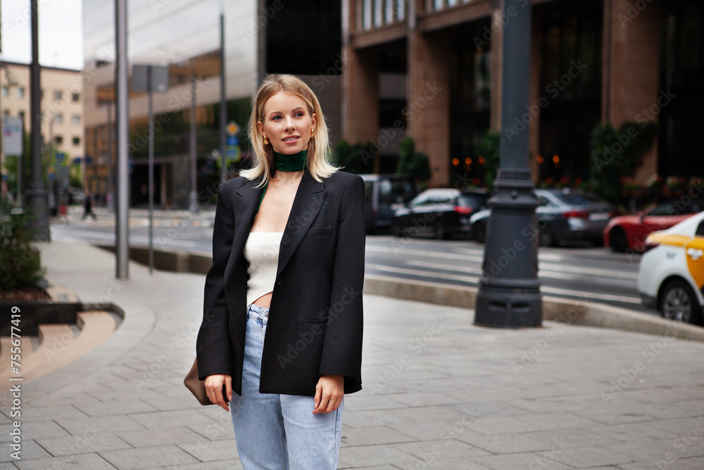 Beautiful young stylish blonde woman on city street against backdrop of building, wearing fashionable clothes, black blazer and silk scarf. Fashion model looks away