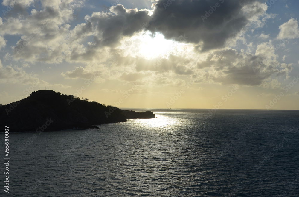 Fototapeta premium Sunset at sea, reflections of sunrays on rippled water surface, clouds, part of an island