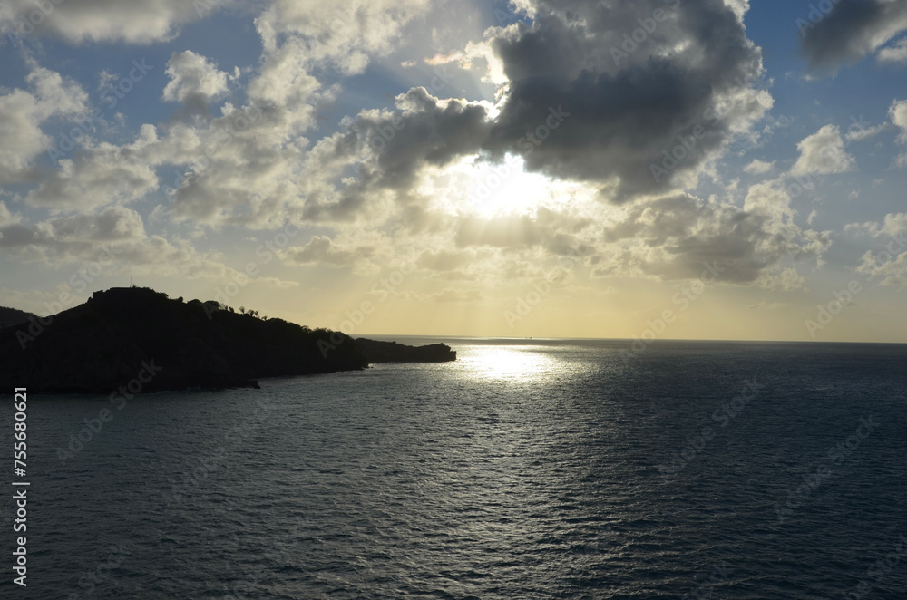 Sunset at sea, reflections of sunrays on rippled water surface, clouds, part of an island Sunset at sea, reflections of sunrays on rippled water surface, clouds, part of an island